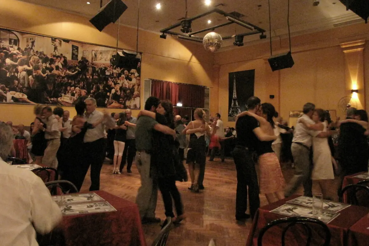 A crowded tango dance hall with couples in close embrace on a wooden floor under a mirror ball