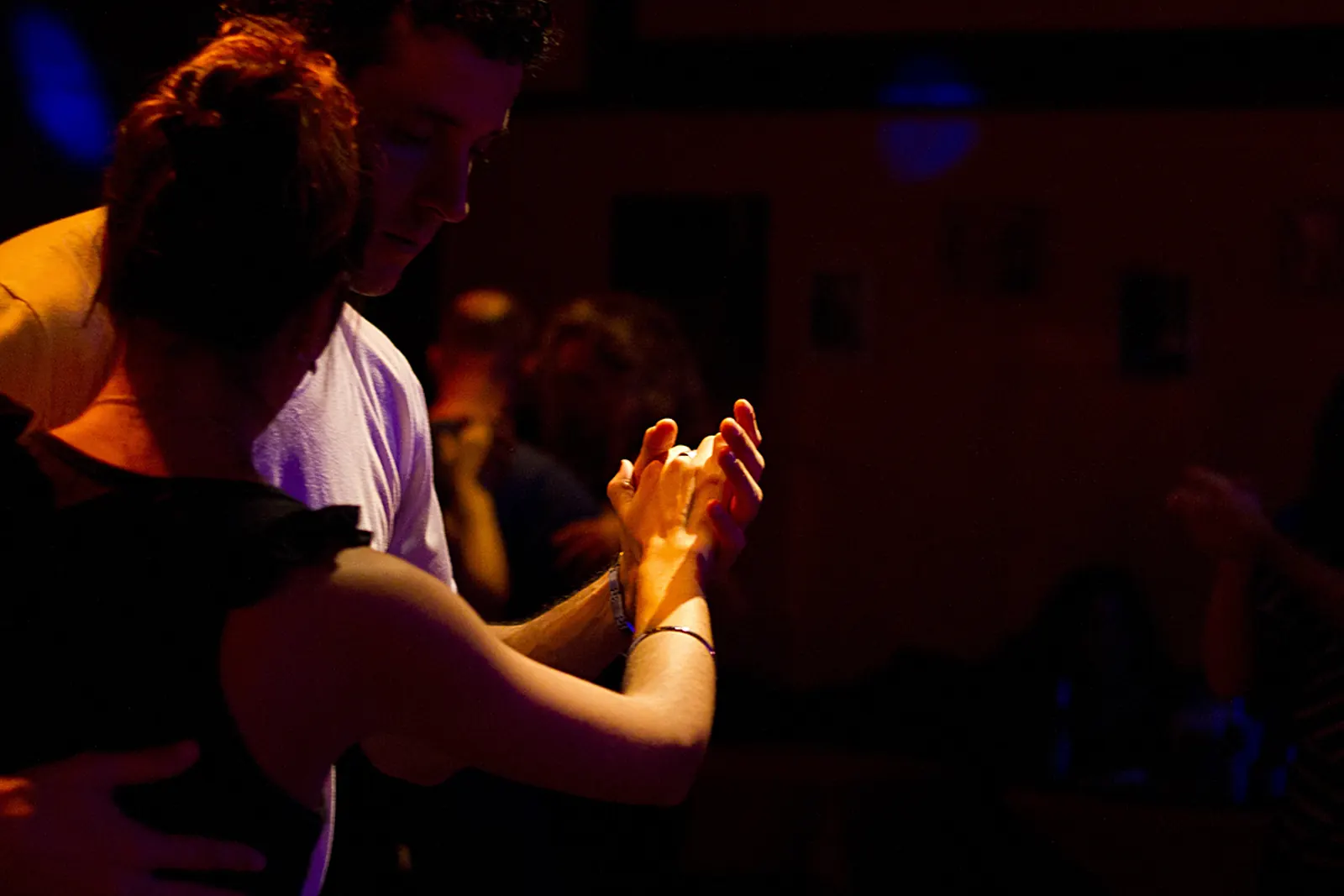 A tango couple's joined hands lit against a dark room, the man's face bowed behind the woman's shoulder
