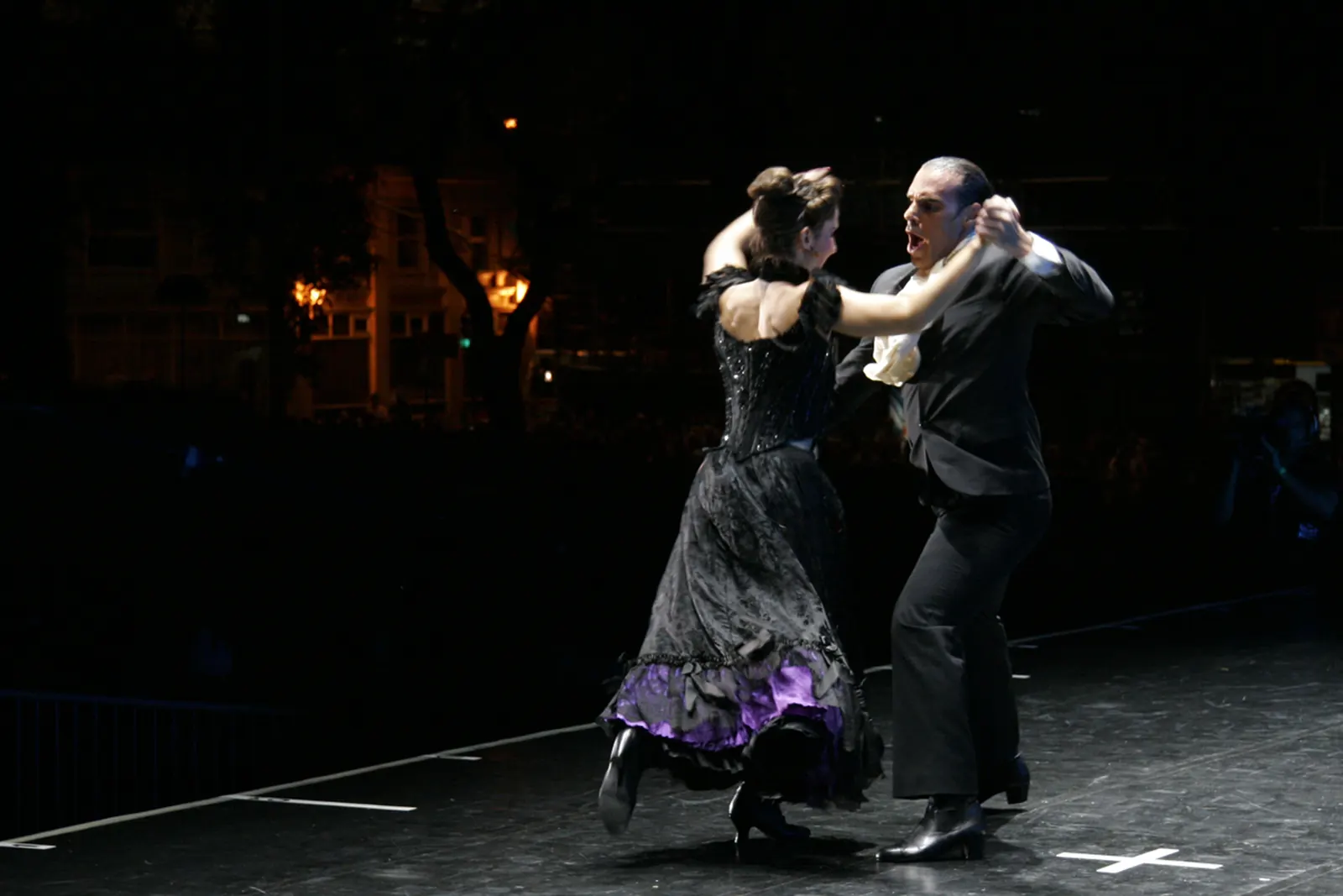 A tango couple in black on a dark stage lit by a single spotlight, the woman's skirt trimmed in purple
